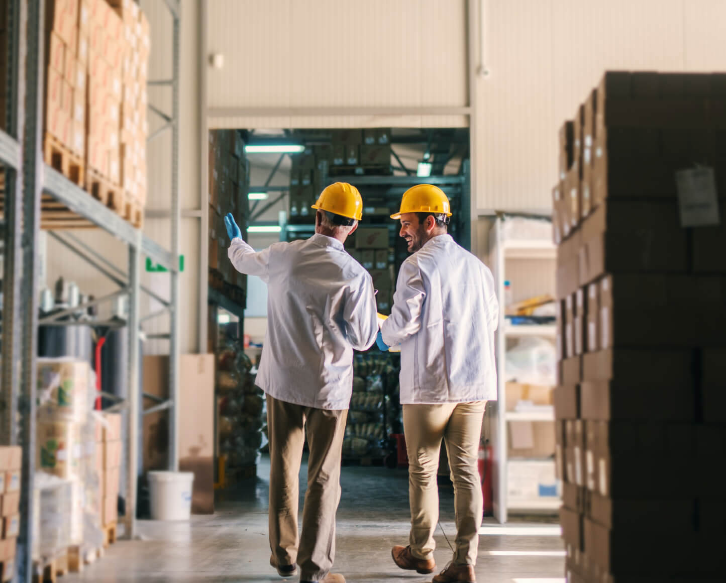 back view of two men wearing labcoats and yellow hardhats pointing at shelves of boxes in a warehouse