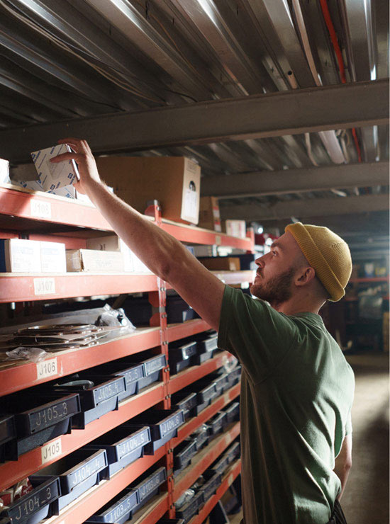 bearded man wearing beanie reaching for small box on top shelf in basement storage aisle