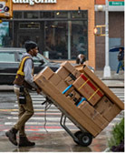 man of color wearing beanie with headphones in pushing stacked boxes on wheeled hand truck on rainy sidewalk in urban area