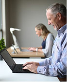man standing at counter smiling at laptop and typing with woman in background blurred and doing the same thing